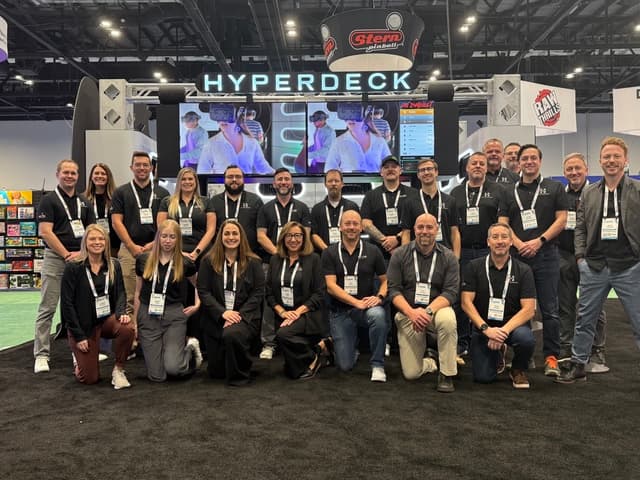 A group of people posing in front of a "HYPERDECK" booth at a convention, wearing name badges and casual attire.