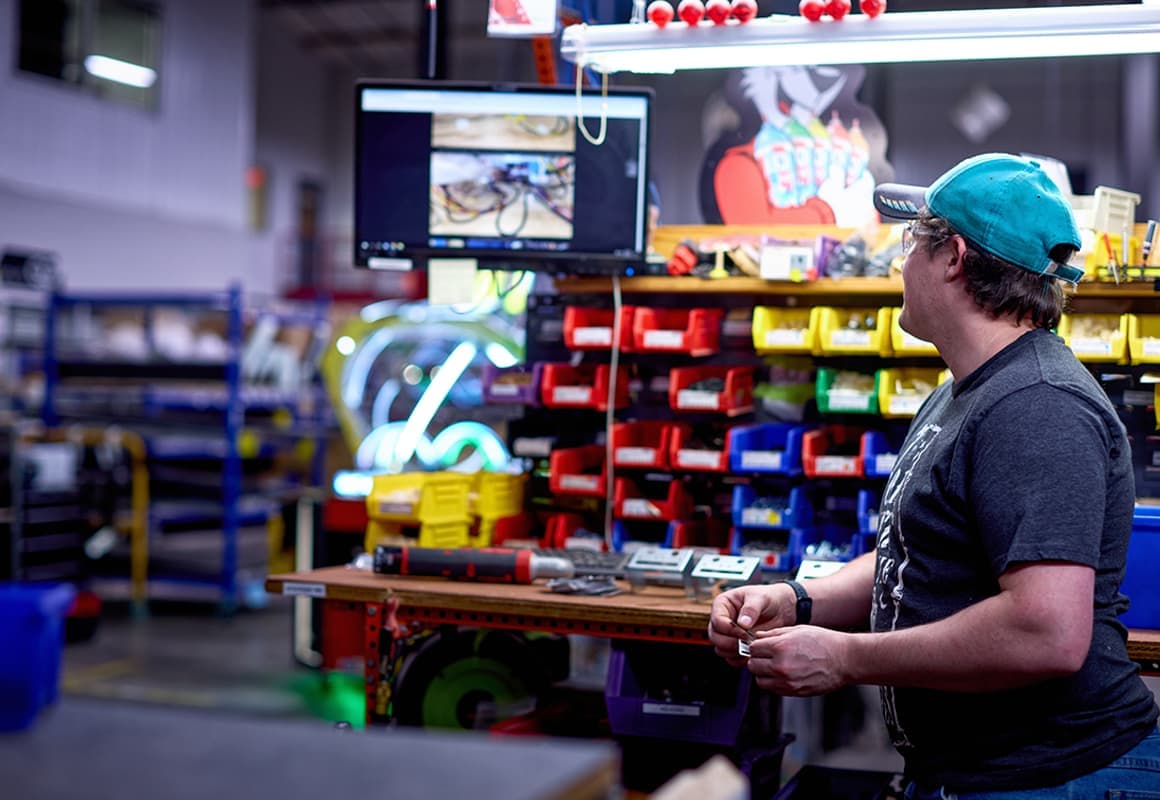 A person in a cap stands in a workshop, looking at a screen displaying an image. Shelves with colorful bins and tools are in the background.